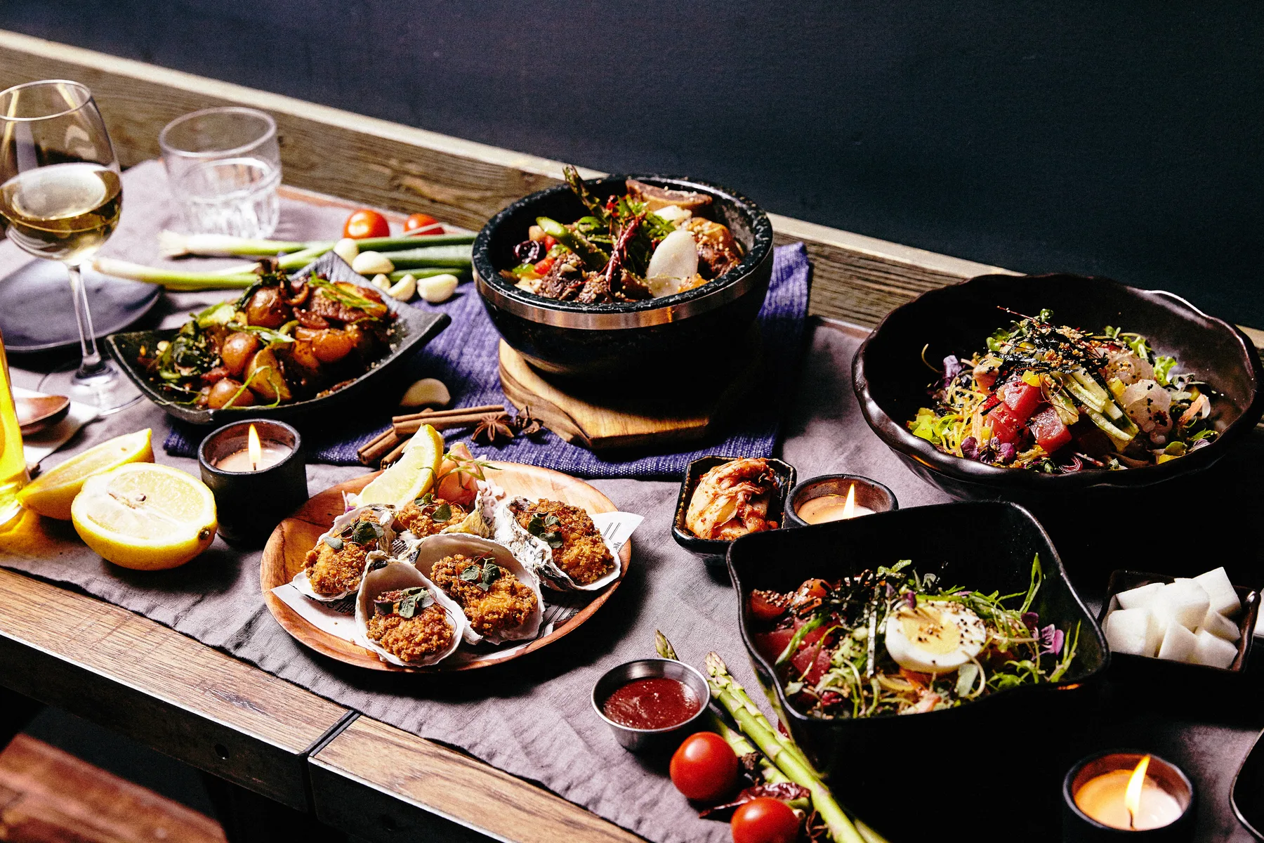 A spread of Korean-inspired dishes on a dark wooden table with a copper plate of scallops